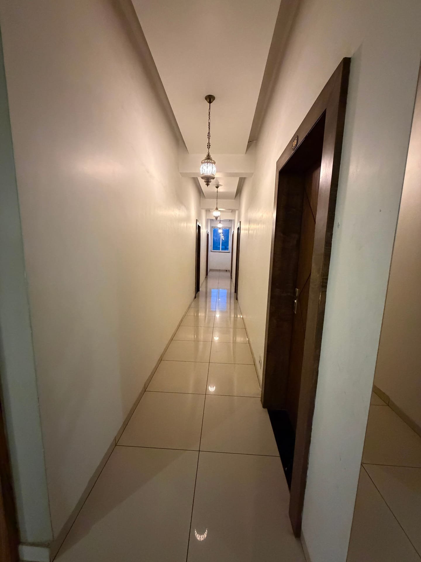 A guest-room corridor with polished cream floor tiles, dark timber doors and hanging brass lanterns leading to a bright window at the end