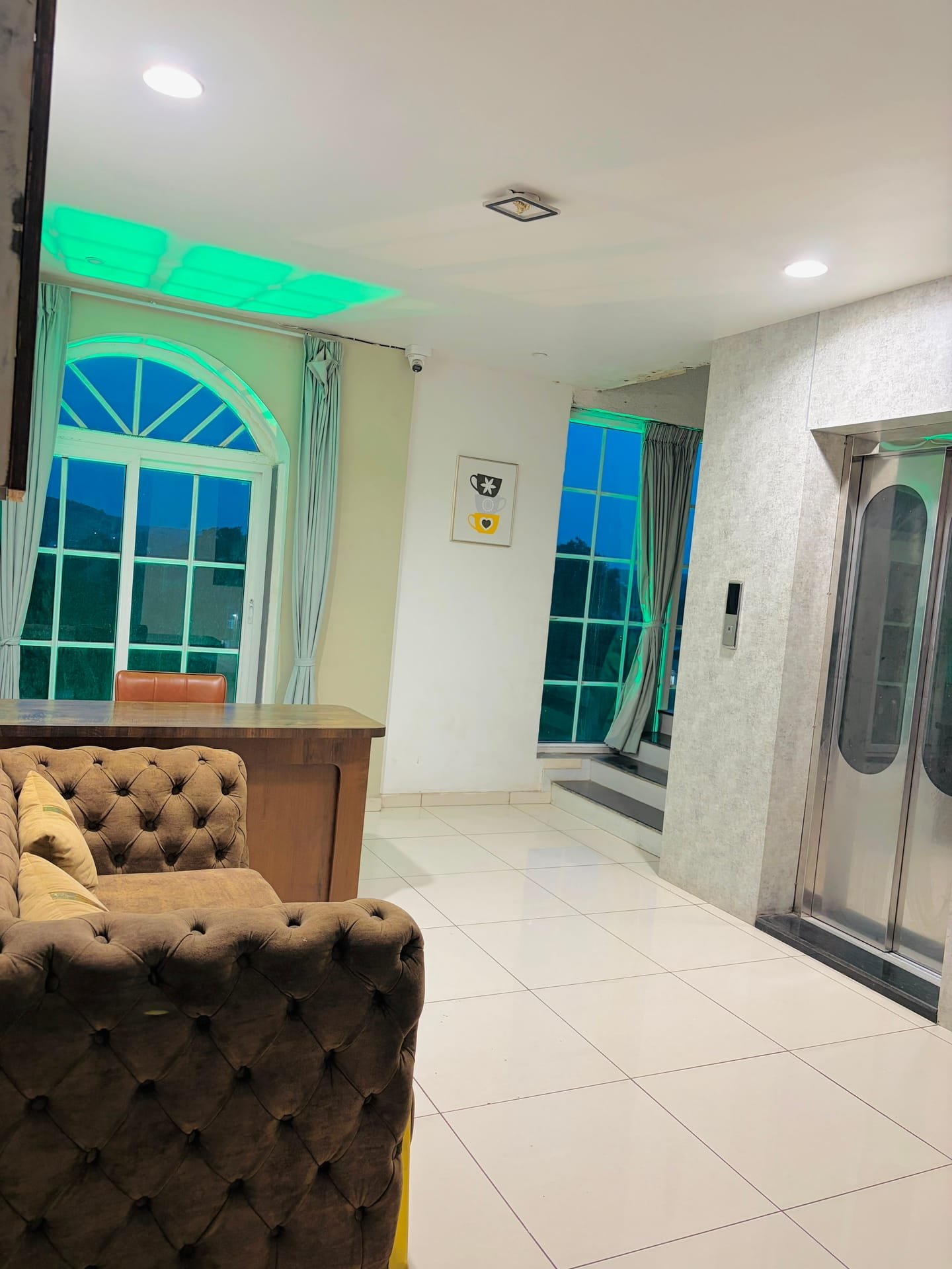 Reception lounge with a tufted chesterfield sofa, a timber counter and the lift beyond, tall windows lit softly in blue and green from cove lighting