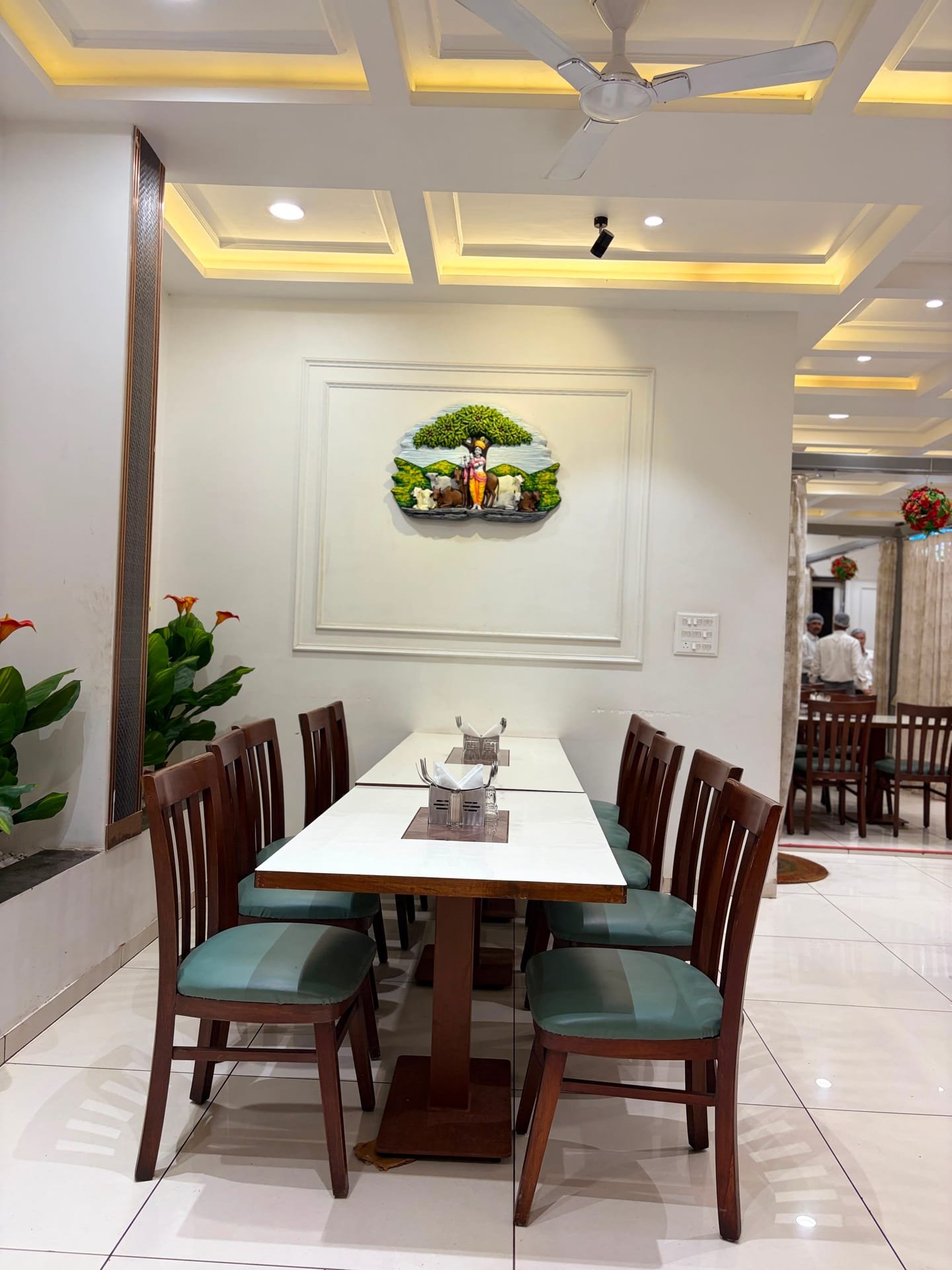A dining table in the restaurant with wooden chairs and sage-green seat cushions, beneath a wall relief of Krishna standing under a tree with cows