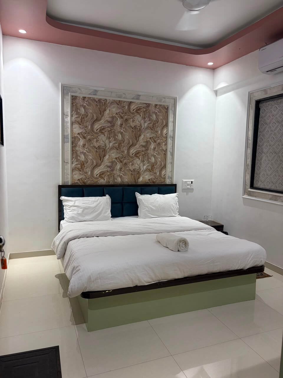 Air-conditioned guest room seen from the door — marble-framed floral feature wall, blue tufted headboard, wide bed in white linen and a sage-green bed base on polished tile