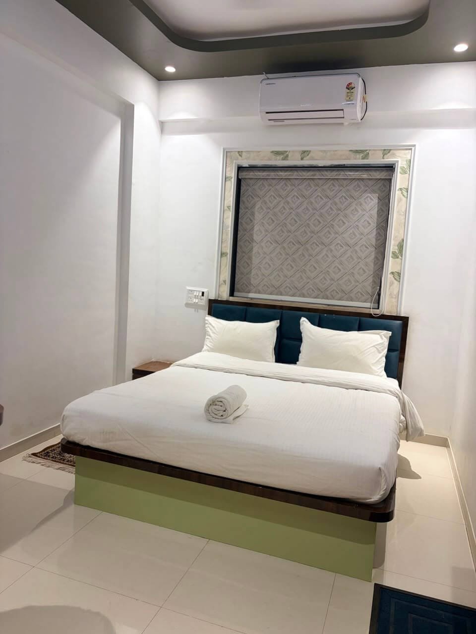 Air-conditioned room with a blind-covered window framed in leaf-patterned border, blue tufted headboard and a rolled towel on the bed