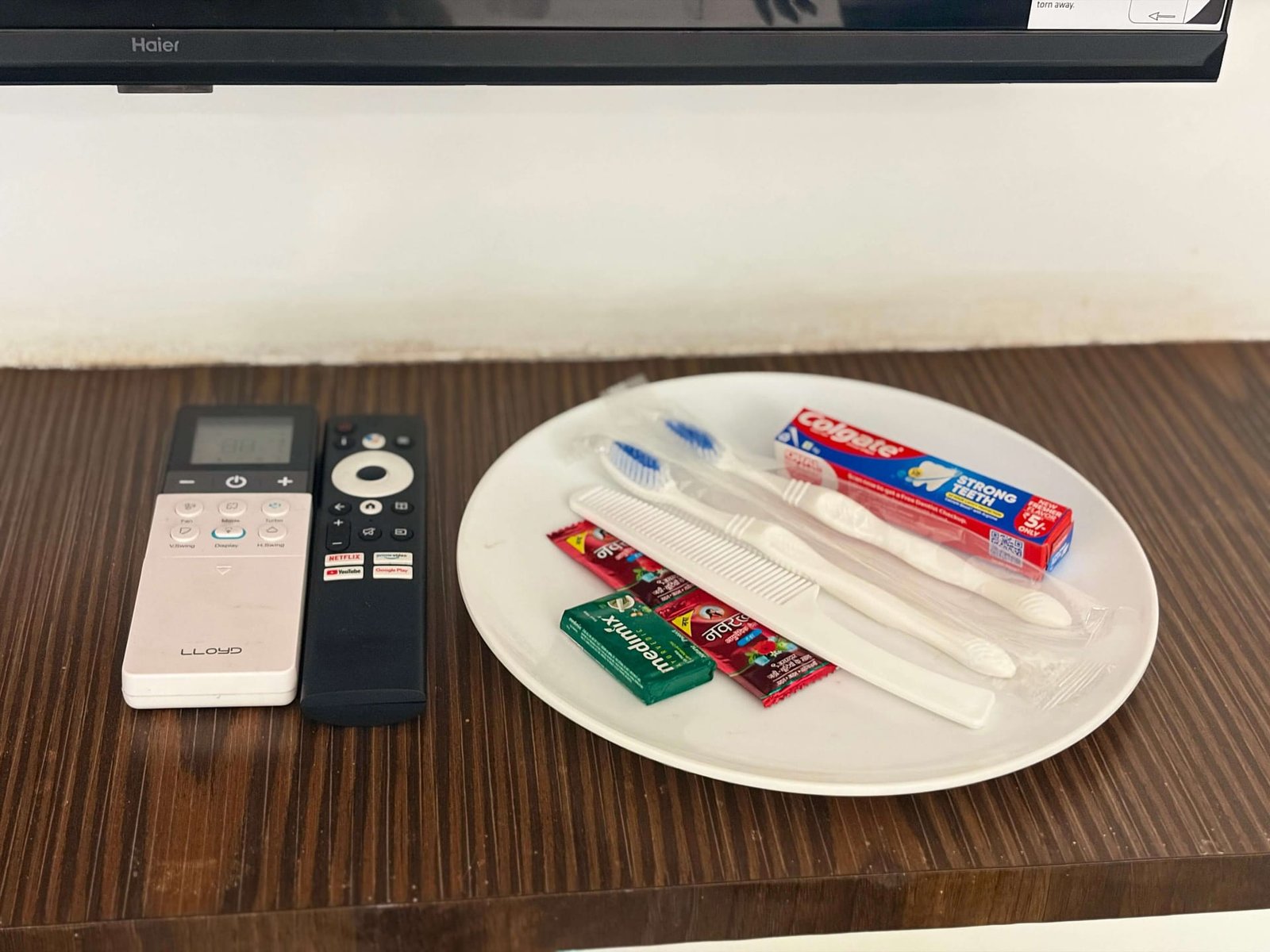 Complimentary in-room amenity set on a plate — toothbrushes, toothpaste, comb and soap — beside the air-conditioner and television remotes