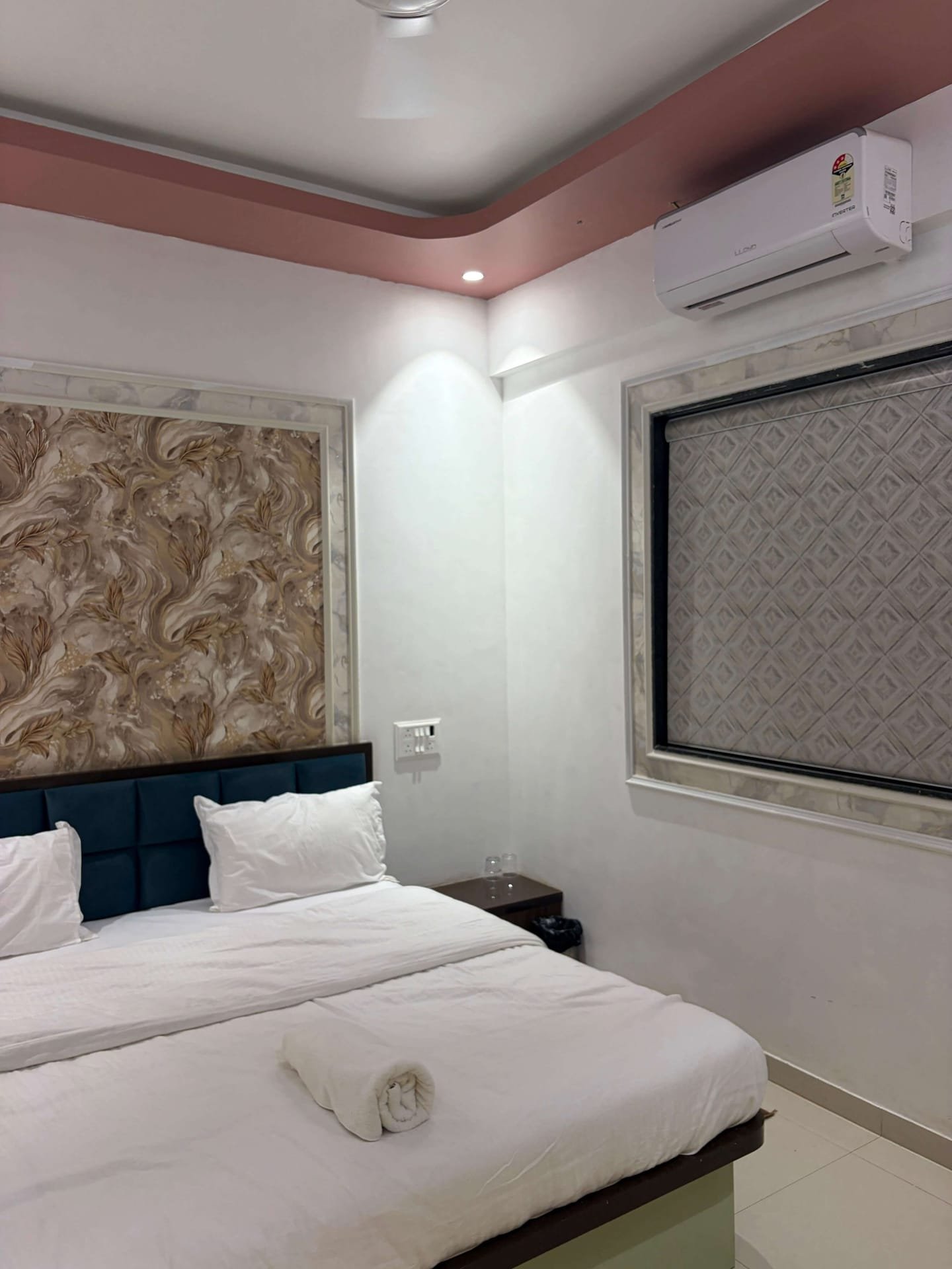 Guest room with a wide bed in crisp white linen, blue upholstered headboard, marble-framed feature wall and a wall-mounted air conditioner