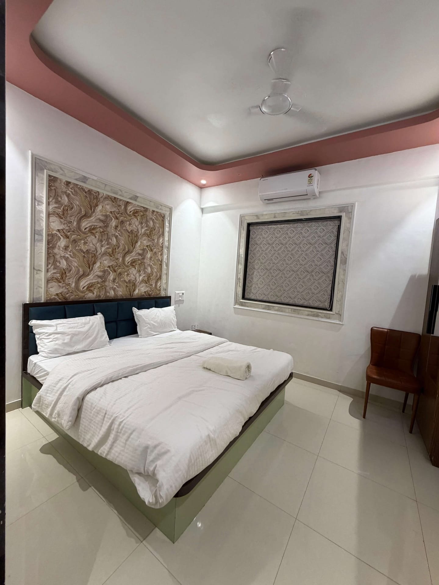 Wide view of a deluxe room showing the bed, patterned feature wall, air conditioning, seating chair and glossy tiled floor
