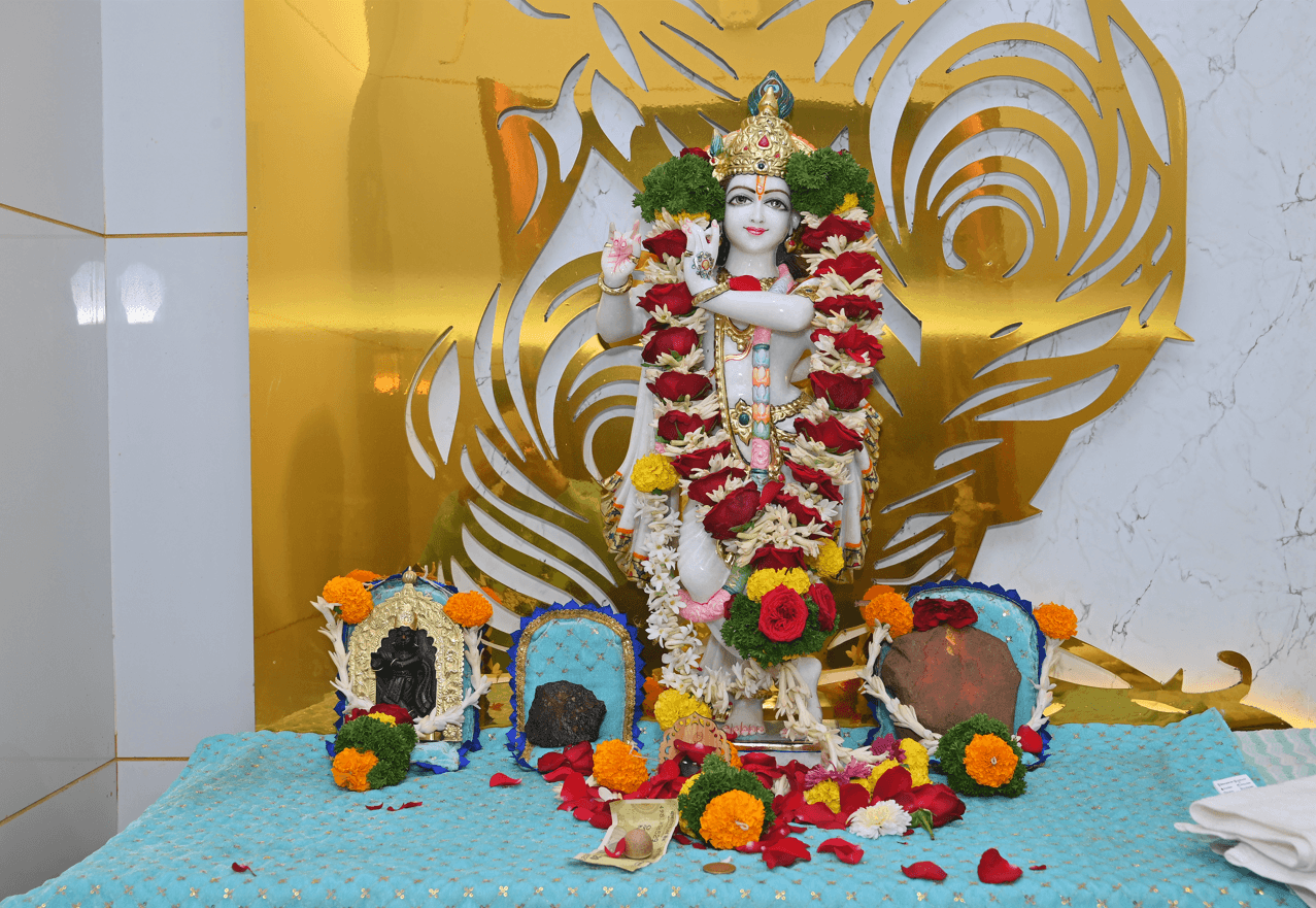 Marble Krishna murti garlanded with roses and marigolds before a gold peacock-feather backdrop, with offerings laid on a blue cloth